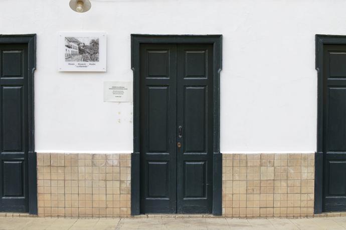 Edificio donde se ven tres puertas de verde oscuro en unas paredes blancas con mosaicos y unas placas con información