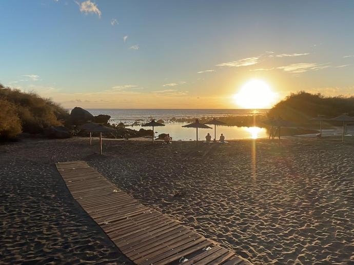 Amplia playa de arena al atardecer que tiene un paseo de tablas de madera y sombrillas en la propia playa